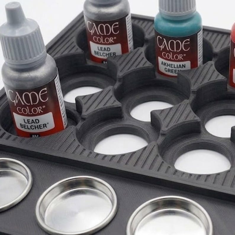 May include: A black paint bottle holder with multiple compartments, holding several bottles of Game Color acrylic paint. The bottles are labeled with paint names like "Lead Belcher" and "Akhelian Green". Several small, round metal containers are in the foreground.