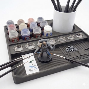 May include: A gray plastic hobby paint station with various paint bottles, small metal mixing wells, and paint brushes. A miniature figurine is positioned in the center. A white ceramic cup holds additional paint brushes.