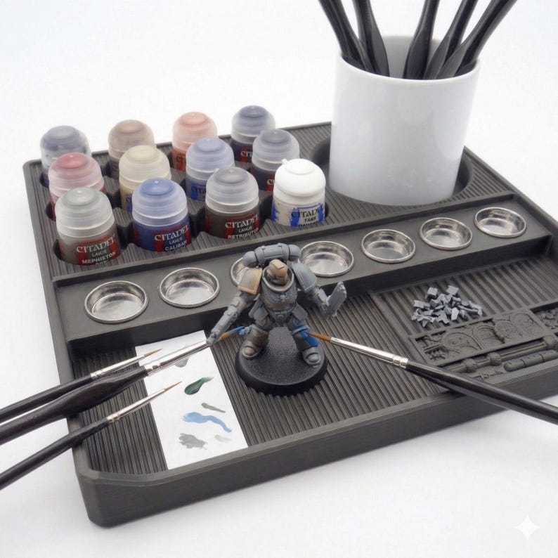 May include: A gray plastic hobby paint station with various paint bottles, small metal mixing wells, and paint brushes. A miniature figurine is positioned in the center. A white ceramic cup holds additional paint brushes.