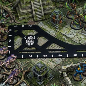 K&ouml;nnte beinhalten: Eine Tabletop-Gaming-Szene mit Miniaturfiguren, W&uuml;rfeln und einem schwarzen Messwerkzeug mit wei&szlig;en Markierungen. Die Szene umfasst bemalte Figuren, ein Ma&szlig;band und eine detaillierte Gel&auml;ndeeinstellung mit Miniaturruinen und -laub.