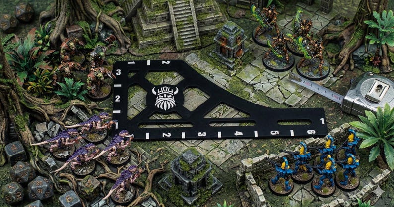 K&ouml;nnte beinhalten: Eine Tabletop-Gaming-Szene mit Miniaturfiguren, W&uuml;rfeln und einem schwarzen Messwerkzeug mit wei&szlig;en Markierungen. Die Szene umfasst bemalte Figuren, ein Ma&szlig;band und eine detaillierte Gel&auml;ndeeinstellung mit Miniaturruinen und -laub.