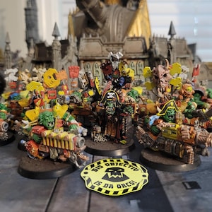 Può includere: Una collezione di miniature di Ork, alcune montate su veicoli, con un Ork a tema pirata al centro. Le figure sono dipinte in vari colori, tra cui verde, giallo e marrone. Un cartello giallo e nero recita "DIS OBJECTIVU IS DA ORCS!"