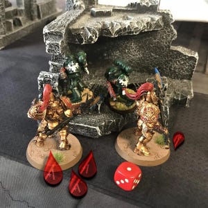 Puede incluir: Primer plano de un juego de guerra de mesa con cuatro figuras en miniatura. Dos figuras están pintadas con armadura dorada y capas rojas, mientras que las otras dos están pintadas con armadura verde. Las figuras están sobre bases circulares. También hay cuatro dados rojos sobre la mesa.