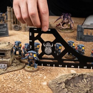 K&ouml;nnte beinhalten: Ein schwarzes, rechtwinkliges Messwerkzeug mit wei&szlig;en Markierungen wird verwendet, um Miniaturfiguren auf einem Tisch zu messen. Die Figuren sind Teil eines Tabletop-Kriegsspiels, mit anderen Spielsteinen und Gel&auml;nde sichtbar.