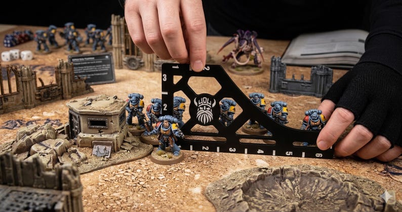 K&ouml;nnte beinhalten: Ein schwarzes, rechtwinkliges Messwerkzeug mit wei&szlig;en Markierungen wird verwendet, um Miniaturfiguren auf einem Tisch zu messen. Die Figuren sind Teil eines Tabletop-Kriegsspiels, mit anderen Spielsteinen und Gel&auml;nde sichtbar.