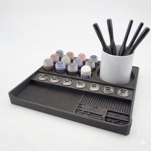 May include: A dark gray paint station with a variety of paint bottles, small metal paint wells, and a white ceramic cup holding black paint stirrers. The station is designed to organize painting supplies.