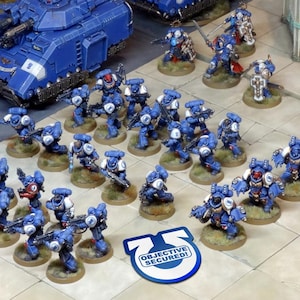 Op de afbeelding: Een tabletop-oorlogsvoeringsscène met een blauw-wit geschilderd leger van miniatuur space marines. De miniaturen zijn in een gevechtsformatie op een beige oppervlak gerangschikt. Een blauw-witte tank bevindt zich op de achtergrond. Een blauwe en witte ronde token met de tekst "OBJECTIVE SECURED!" bevindt zich op de voorgrond.