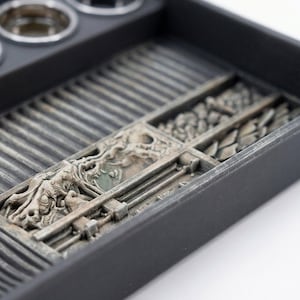 May include: A black organizer tray with compartments and a detailed, textured design. The tray features a series of rectangular sections with various patterns and a section with a detailed relief carving. The tray is designed to hold small items.