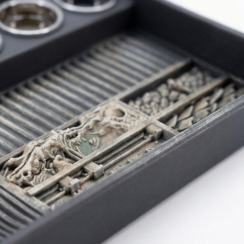May include: A black organizer tray with compartments and a detailed, textured design. The tray features a series of rectangular sections with various patterns and a section with a detailed relief carving. The tray is designed to hold small items.