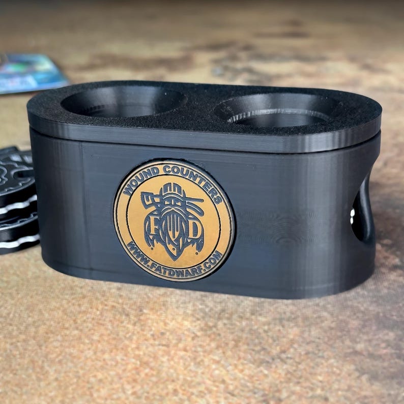 May include: Black plastic wound counter holder with two slots for dice. The holder has a gold logo that says "Wound Counters" with a graphic of a skull and crossbones and the website "www.fatdwarf.com".