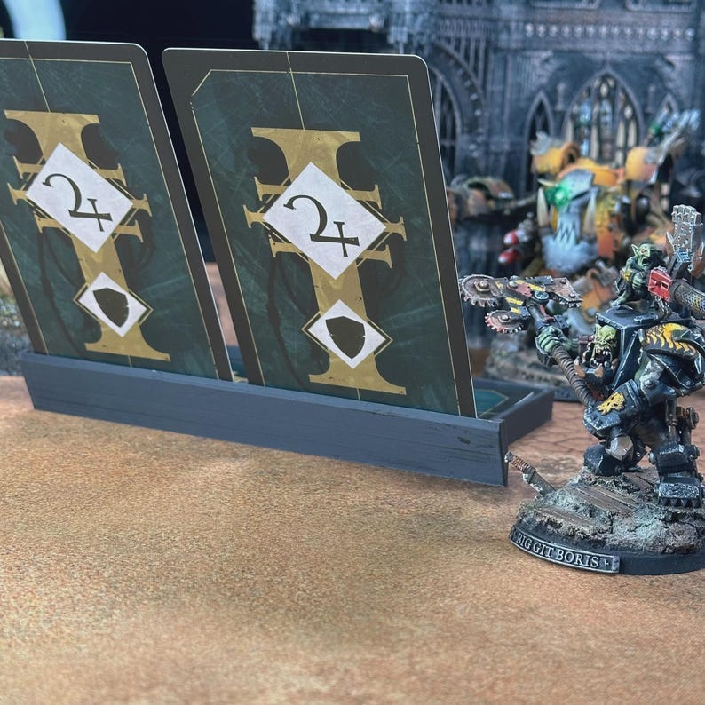May include: A dark gray plastic card holder containing two rectangular game cards with dark green backgrounds, gold accents, and white diamond symbols featuring the number two and a shield icon, positioned next to a painted miniature figure.