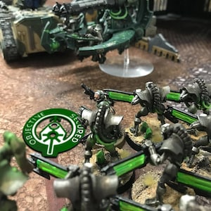 May include: A close-up of a group of miniature figures, painted in green and silver, holding weapons. The figures are standing on a brown surface with green and brown accents. A green circular token with the words "Objective Secured" is in the foreground.