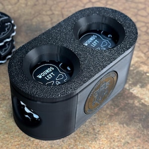 May include: Black plastic container with two circular compartments for holding game pieces. Each compartment has a black and white skull graphic with the text "Wounds Left". The container has a gold metal emblem with a design of a bird and the text "Board Game Universe".