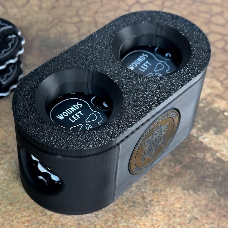 May include: Black plastic container with two circular compartments for holding game pieces. Each compartment has a black and white skull graphic with the text "Wounds Left". The container has a gold metal emblem with a design of a bird and the text "Board Game Universe".