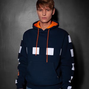Könnte beinhalten: Ein marineblauer Kapuzenpullover mit weißen und orangen Akzenten. Der Pullover hat ein weißes Karomuster an den Ärmeln und ein weißes Karomusterband über der Brust. Die Kapuze ist orange gefüttert.