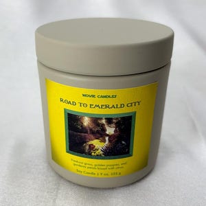 May include: A cylindrical, light gray candle with a matching lid. The candle has a yellow label with the text "ROAD TO EMERALD CITY" and an image of a forest scene. The label also states the candle is 9 oz, 255 g.