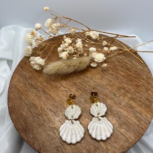Peut inclure: Une paire de boucles d'oreilles pendantes composées de deux breloques superposées en forme de coquillage blanc irisé, suspendues à des tiges en métal doré. Elles sont présentées avec des fleurs séchées blanches et un plumeau d'herbe séchée.