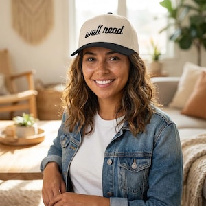 May include: A beige baseball cap with a black peak and the words "well read" in black script. The hat is worn by a person wearing a denim jacket and a white t-shirt.