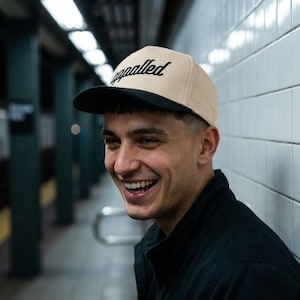 May include: A beige and black baseball cap with the word "appalled" in black script. The cap has a flat brim and is worn by a person smiling in a subway station.