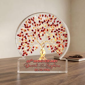 Könnte beinhalten: Eine klare Acrylbaumskulptur mit kreisförmigem Design, mit roten und goldenen blattförmigen Akzenten. Der Sockel zeigt den Text "40. JAHRESTAG" und die Namen "David & Sophia" mit dem Datum "05.12.1986".
