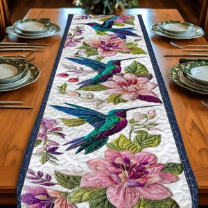 May include: A white quilted table runner featuring a vibrant design of hummingbirds and flowers. The hummingbirds have teal, purple, and green feathers. The flowers are pink and purple with green leaves. The runner has a navy blue border.