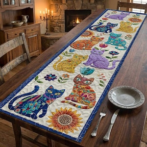 Może przedstawiać: Kremowy bieżnik na stół z kolorowymi wzorami kotów. Koty są przedstawione w różnych pozach i kolorach, otoczone kwiatami i słonecznikami. Bieżnik ma niebieską obwódkę i leży na drewnianym stole.