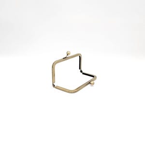 May include: A bronze-coloured, rectangular purse frame. The frame is open, showing the interior. It features a ball clasp on each side and a decorative ball at the top. Suitable for crafting projects.