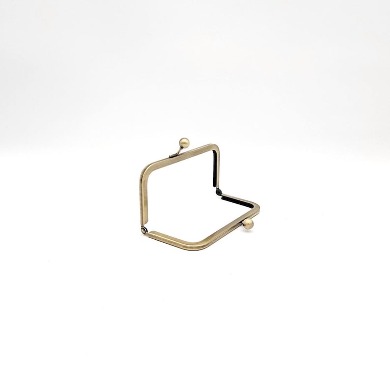 May include: A bronze-coloured, rectangular purse frame. The frame is open, showing the interior. It features a ball clasp on each side and a decorative ball at the top. Suitable for crafting projects.