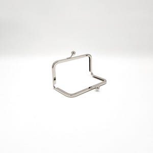 May include: A silver metal purse frame, rectangular with rounded corners. It features a hinged opening and a small ball closure on each side. Ideal for crafting handmade purses or bags. The frame is made of a shiny, metallic material.