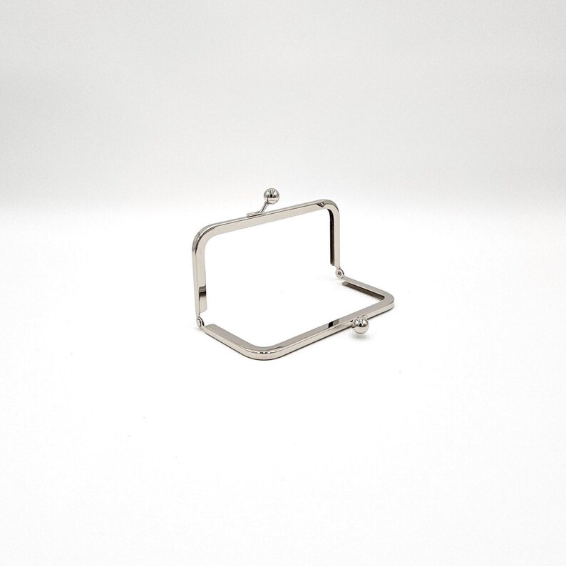 May include: A silver metal purse frame, rectangular with rounded corners. It features a hinged opening and a small ball closure on each side. Ideal for crafting handmade purses or bags. The frame is made of a shiny, metallic material.