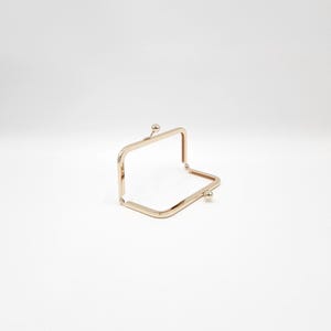 May include: A gold-coloured metal purse frame, rectangular with rounded corners. It features a ball clasp on each side and is open, showing the interior. The frame is designed for attaching to a purse or bag.