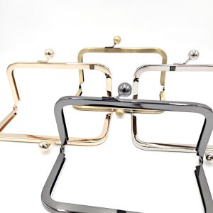 May include: Close-up of several metal purse frames in gold, silver, and gunmetal finishes. Each frame has a rectangular shape with a ball clasp closure. The frames are displayed against a white background.