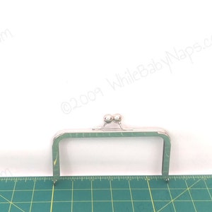 May include: A silver-coloured rectangular purse frame with a ball clasp. The frame has a rectangular shape with rounded corners and pointed ends. The frame is placed on a green cutting mat with a ruler.