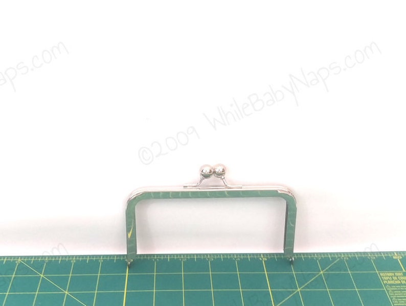 May include: A silver-coloured rectangular purse frame with a ball clasp. The frame has a rectangular shape with rounded corners and pointed ends. The frame is placed on a green cutting mat with a ruler.