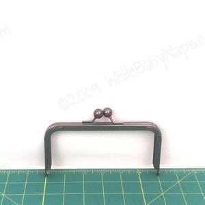 May include: A dark grey metal purse frame, rectangular with two small ball closures. It sits on a green cutting mat with measurements. The background is white. The frame's dimensions are in centimetres.