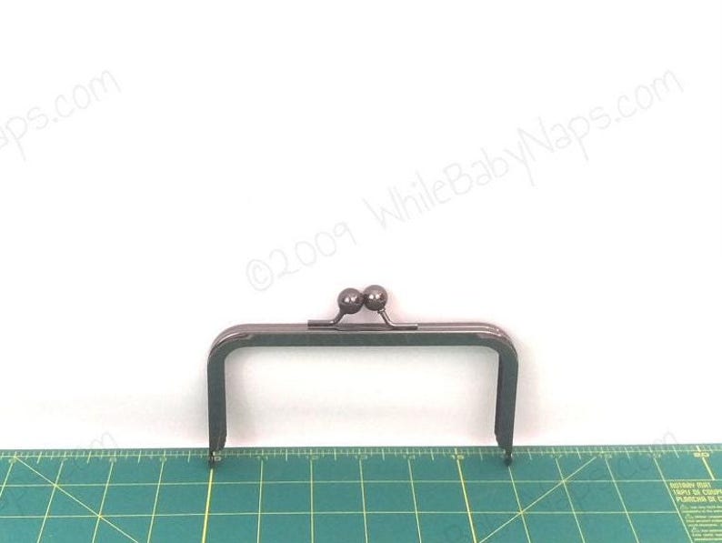 May include: A dark grey metal purse frame, rectangular with two small ball closures. It sits on a green cutting mat with measurements. The background is white. The frame's dimensions are in centimetres.