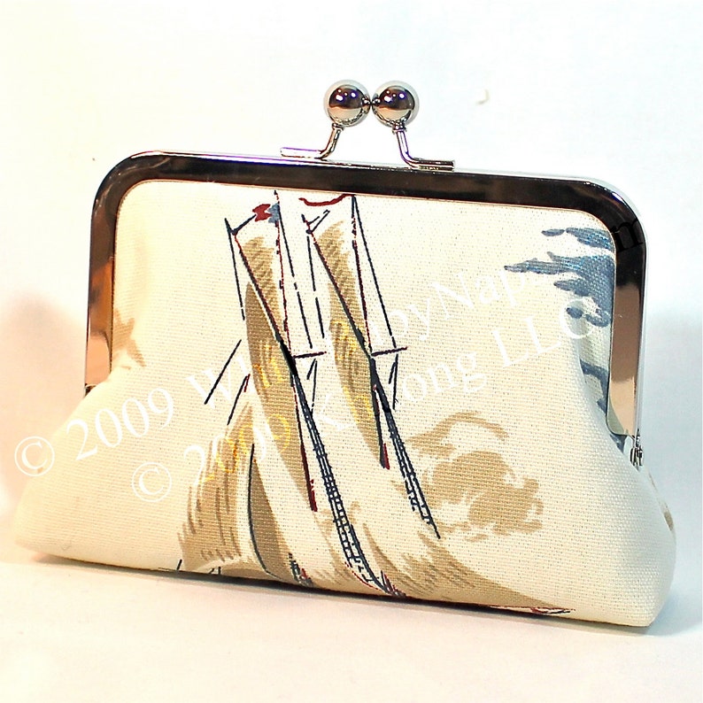 May include: Cream-coloured clutch bag with a sailboat pattern. The fabric features a nautical design with sailboats in beige, blue, and red. The frame is silver-toned with a kiss-lock closure.