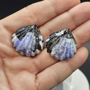 May include: Two seashell-shaped earrings. Each earring features a black, white, and blue design. The earrings are held in a hand, showcasing their intricate details and unique shape. The earrings have a glossy finish.
