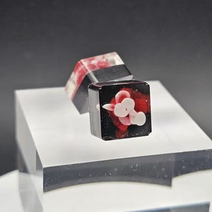 May include: Two square resin art pieces. One is black with a red and white floral design, the other is black with red and white accents. Both are displayed on a clear acrylic block against a dark background.