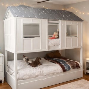 May include: A white bunk bed with a house-shaped frame and a gray roof. The bed has two levels, with windows and a pull-out trundle. Soft toys and bedding are visible on the beds. The room is lit with string lights.