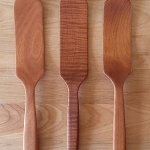 May include: Three wooden spatulas with a natural finish are displayed on a wooden surface. The spatulas vary slightly in shape and wood grain, showcasing the natural beauty of the wood. The spatulas are ideal for cooking and baking.