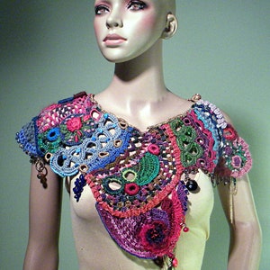 May include: A colourful crocheted collar with beads and fringe. The collar is made of various shades of pink, red, blue, green, and brown yarn.
