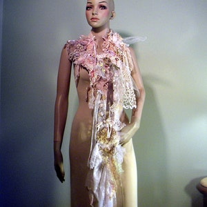May include: A decorative scarf with a layered design, featuring lace, fabric flowers, and ribbons in shades of pink, cream, and gold. The scarf is displayed on a mannequin, showcasing its intricate details and textures. The scarf is a unique accessory.