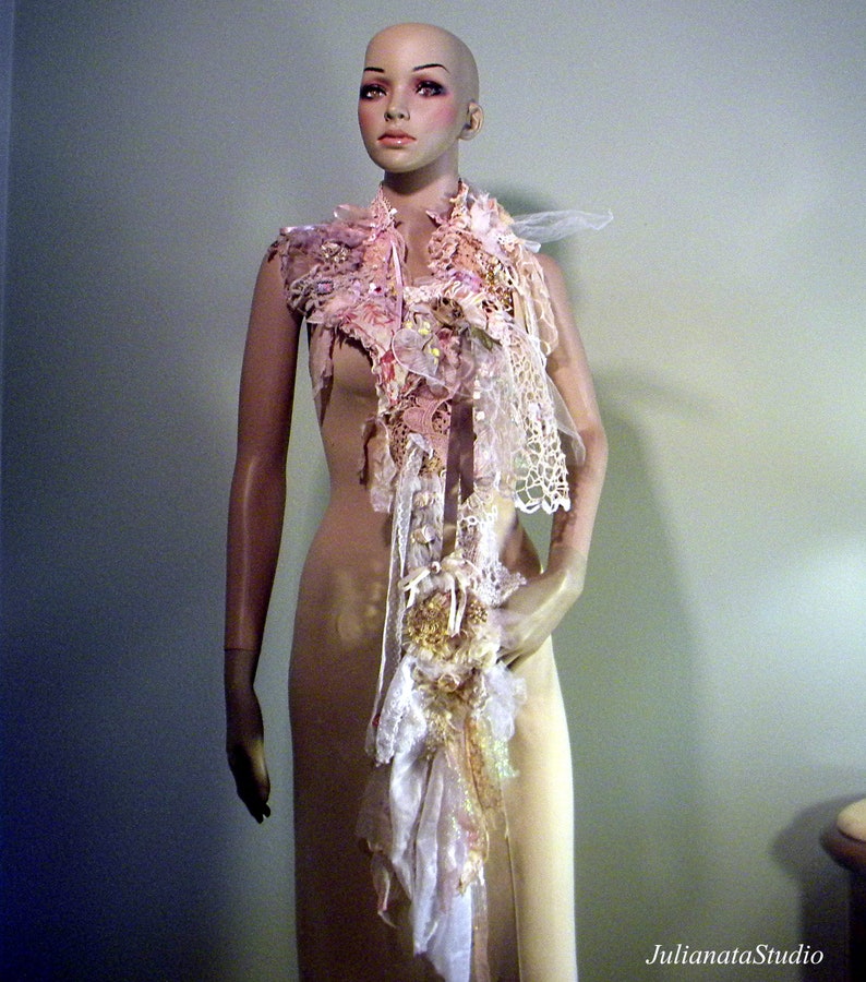 May include: A decorative scarf with a layered design, featuring lace, fabric flowers, and ribbons in shades of pink, cream, and gold. The scarf is displayed on a mannequin, showcasing its intricate details and textures. The scarf is a unique accessory.