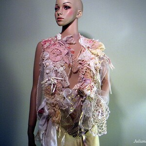 May include: A handmade, layered vest with a romantic, bohemian aesthetic. The vest features a mix of lace, fabric scraps, and embellishments in shades of pink, cream, and gold. Delicate ribbons and floral accents add to the intricate design. The vest is displayed on a mannequin.