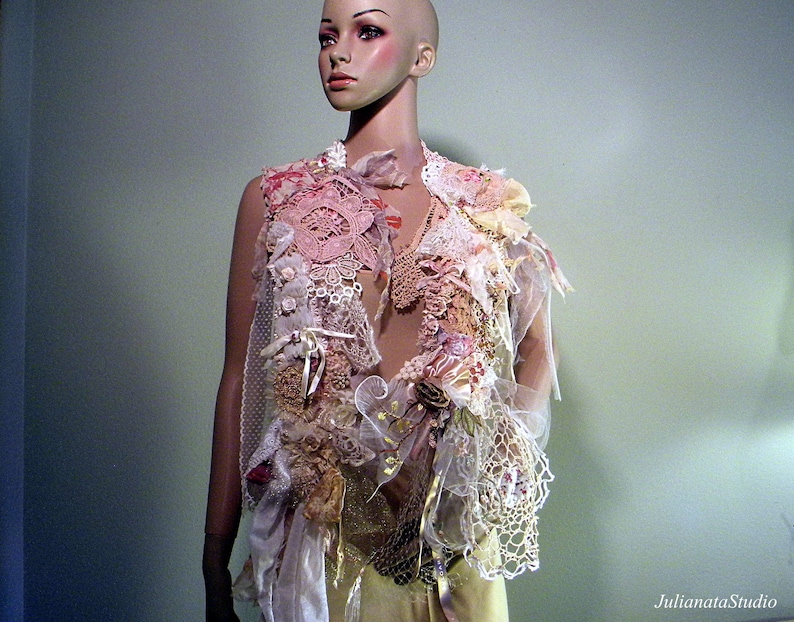 May include: A handmade, layered vest with a romantic, bohemian aesthetic. The vest features a mix of lace, fabric scraps, and embellishments in shades of pink, cream, and gold. Delicate ribbons and floral accents add to the intricate design. The vest is displayed on a mannequin.