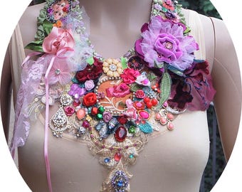 Puede incluir: Un collar llamativo y colorido con una variedad de texturas y adornos, incluyendo flores, cuentas y encaje. El collar es una mezcla de tonos rosas, rojos, verdes y dorados.
