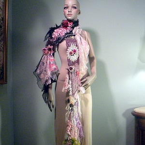 Könnte beinhalten: Dekorativer Schal mit floralen und Spitzenakzenten, präsentiert auf einer Schaufensterpuppe. Der Schal weist eine Mischung aus rosa, lila und schwarzen Stoffen auf, mit einem langen, fließenden Design. Die Schaufensterpuppe trägt ein cremefarbenes Kleid.