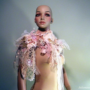 May include: A decorative shoulder piece on a mannequin. The piece features layers of lace, fabric, and floral embellishments in shades of pink, cream, and white. The mannequin has a neutral skin tone and is set against a soft green background.
