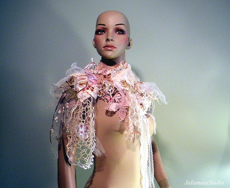 May include: A decorative shoulder piece on a mannequin. The piece features layers of lace, fabric, and floral embellishments in shades of pink, cream, and white. The mannequin has a neutral skin tone and is set against a soft green background.